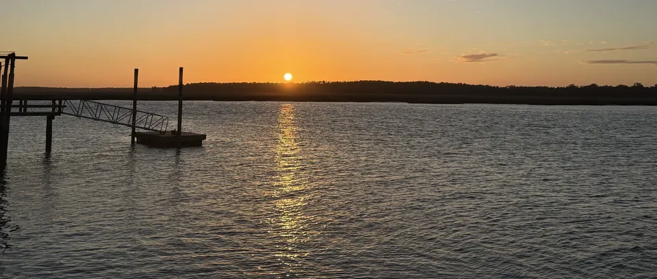 Sun setting beyond the marsh across the Sapelo River with a golden reflection on the water and a dock in the foreground.